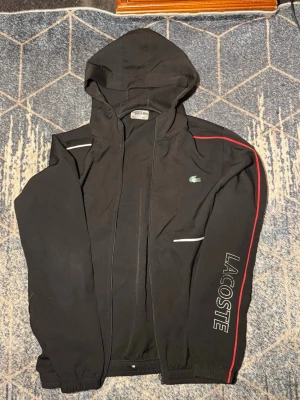 Svart hoodie från Lacoste med röd detalj - Snygg svart hoodie från Lacoste med dragkedja, huva och elastiska muddar. Har en röd linje längs ärmen och reflekterande detaljer. Stort Lacoste-tryck på ena ärmen och klassiska krokodilloggan på bröstet. Perfekt för en sportig och trendig look.