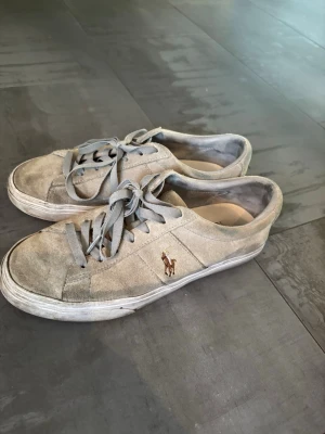 Gråa sneakers från Polo Ralph Lauren - Gråa sneakers i mocka från Polo Ralph Lauren med klassisk låg profil och snörning. Ikonisk broderad logga på sidan och vit sula. Perfekta för en avslappnad streetwear-look.