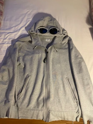 Grå hoodie med glasögon från C.P. Company - Säljer en grå hoodie från C.P. Company. Hoodien har dragkedja framtill, två sidofickor med dragkedja och ribbade muddar.