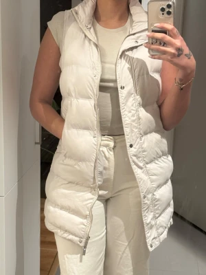Vit lång dunväst med huva - Säljer en snygg och trendig vit dunväst med huva och dragkedja framtill. Västen är quiltad, har tryckknappar och två sidofickor. Perfekt för lager på lager och ger en clean look till din outfit. Passar dig som gillar stilrena och bekväma plagg.