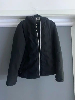 Svart pufferjacka med huva - Snygg svart pufferjacka med huva och dragkedja framtill. Jackan har en enkel, stilren design och är vadderad för extra värme. Passar perfekt för kalla vinterdagar och har två sidofickor. Materialet ser ut att vara syntetiskt och jackan har en rak passform.