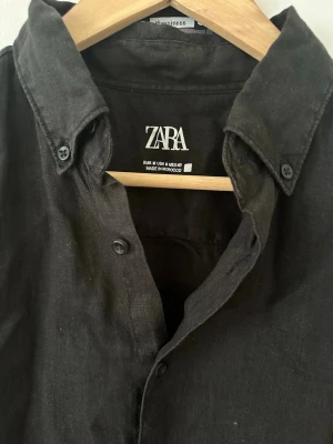 Svart linneskjorta från Zara - Stilren svart skjorta från Zara i 100% linne. Skjortan har klassisk krage, knappar framtill och långa ärmar med knapp vid manschetten. Perfekt för en avslappnad men ändå snygg look.