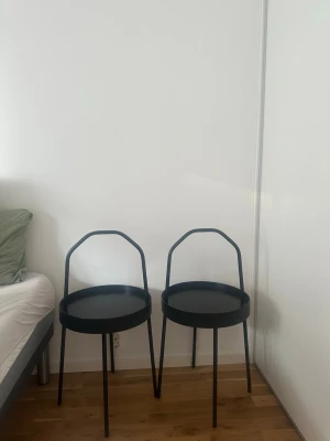 Två svarta sidobord i metall - Säljer två stilrena svarta sidobord i metall med rund form och tre ben. Bordens design har en hög kant runt bordsskivan och ett handtagliknande ryggstöd, vilket ger en modern och unik look. Perfekta som nattduksbord eller små avlastningsbord. (2st =425sek )