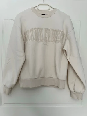Beige sweatshirt Grand Canyon - Beige sweatshirt med broderad text 'Grand Canyon Arizona' på bröstet. Tröjan har rund hals, ribbade muddar vid ärmslut och nederkant samt en avslappnad passform. Perfekt för en casual och trendig look. Använd fåtal gång! Passar även XS