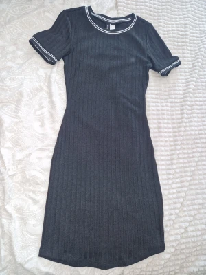 Svart ribbad kortklänning HM divided  - En svart ribbad kortklänning från HM divided med rund halsringning och vita kontrastsömmar vid hals och ärmslut. Klänningen har korta ärmar och en figurnära passform som ger en sportig och stilren look. Jag är 162 & den går ungefär till knäna på mig💥