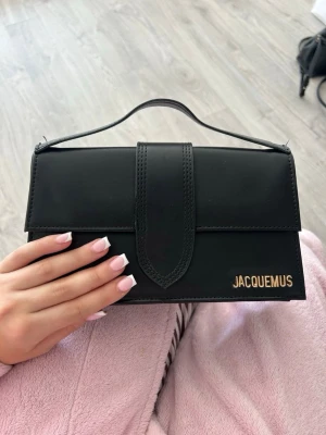 Svart handväska från Jacquemus - Snygg svart handväska från Jacquemus i slätt läder med guldfärgad logga på framsidan. Väskan har ett stilrent lock, topphandtag och en praktisk ficka på baksidan. Perfekt accessoar för att lyfta din outfit.
