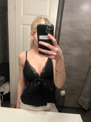 Svart spets linne med rosett - Snygg svart linne i spets med tunna axelband och djup V-ringning. Spetsdetaljer över bysten och midjan samt en liten rosett framtill. Transparenta partier ger en feminin look. Perfekt för dig som vill ha något extra till underkläderna.