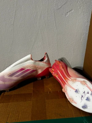 Adidas vita och rosa fotbollsskor - Säljer ett par stilrena fotbollsskor från Adidas i vitt och rosa med lila detaljer. Skorna har en strömlinjeformad design utan snörning och en lätt ovandel i syntetmaterial. Sulan har fasta dobbar för spel på naturgräs. Perfekta för dig som vill sticka ut på planen.