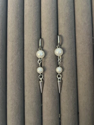 Silvriga örhängen med pärlor och spikes - Säljer ett par stilrena örhängen i silverfärg med två vita pärlor på varje örhänge och en spetsig spike-detalj längst ner. Örhängena har en modern och edgy look som passar perfekt till dig som gillar att sticka ut. Lätta att bära och snygga till alla outfits.