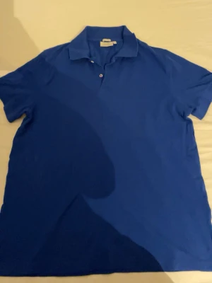 Blå pikétröja med krage - En klassisk blå pikétröja med kort ärm och krage. Tröjan har två knappar vid halsen och är tillverkad i ett pikétyg som känns mjukt och luftigt. Perfekt för en avslappnad och stilren look.