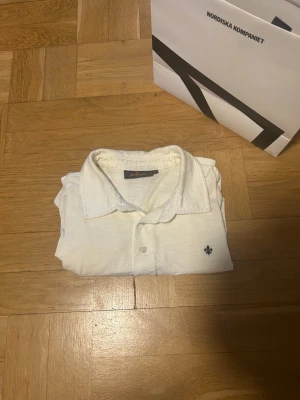 Vit piké från Morris - Vit pikétröja från Morris med klassisk krage och knappslå framtill. Tröjan har korta ärmar och en liten broderad logotyp på bröstet. Tillverkad i mjukt bomullsmaterial som känns skönt mot huden. Påsen ingår ej vid köpet