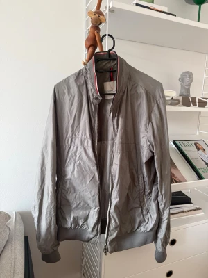 Grå vindjacka från Moncler - Snygg grå vindjacka från Moncler med dragkedja framtill och ribbade muddar vid ärmslut och nederkant. Jackan har en minimalistisk design, ståkrage och tunnt, lätt material som passar perfekt till vår och höst. Klassisk och stilren look med diskret Moncler-logga i nacken. Liter märke vid fickan som ni ser på bilden