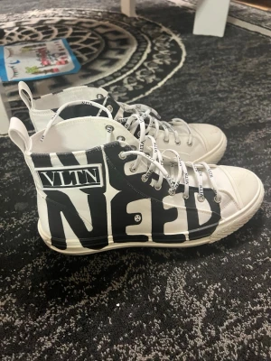 Vita VLTN high top sneakers - Snygga vita high top sneakers från Valentino med svart grafiskt tryck och texten VLTN på sidan. Skorna har vita snören med VLTN-logga och en klassisk gummisula. Tillverkade i canvasmaterial och har en cool streetwear-vibe.