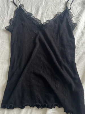 Svart spetslinnetopp från Amisu - Säljer en svart ribbad linnetopp från Amisu med tunna axelband och spetsdetaljer vid urringningen. Toppen är ärmlös och har en lätt vågig kant nertill. Perfekt att styla med jeans eller kjol för en trendig look.