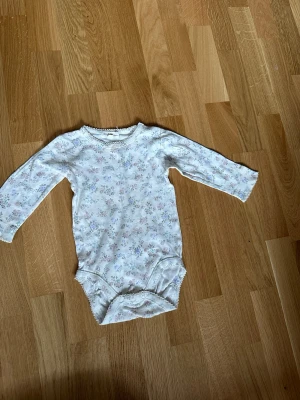 Vit blommig body med lång ärm - Söt vit body med små blommor i pastellfärger och lång ärm. Bodyn har rund halsringning med dekorativ kant och tryckknappar i grenen för enkel påklädning. Perfekt för en mjuk och bekväm känsla hela dagen.