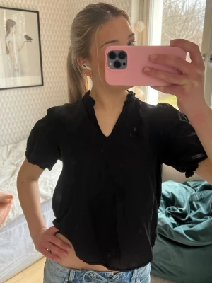 Svart blus med v-ringning - Säljer en svart blus med v-ringning och korta puffärmar. Blusen har en lätt rynkad kant vid halsen och är tillverkad i ett mjukt, tunt tyg som ger en avslappnad passform. Perfekt att styla med jeans eller kjol.