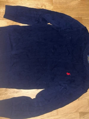 Mörkblå kabelstickad tröja Polo Ralph Lauren - Kabelstickad mörkblå tröja från Polo Ralph Lauren med klassisk röd logga broderad på bröstet. Tröjan har rund halsringning, ribbade muddar och är tillverkad i mjukt material. Perfekt för en stilren och avslappnad look. Storlek S.