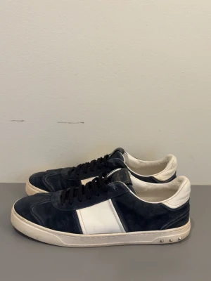 Valentino Flycrew - Snygga mörkblå sneakers från Valentino med vita detaljer och svarta skosnören. Skorna har en låg silhuett, vit sula och coola nitar på hälen. Ovandelen är i mocka och läder vilket ger en lyxig känsla. Perfekta för dig som vill ha en stilren men edgy look.