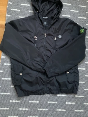Svart vindjacka från Stone Island - Snygg svart vindjacka från Stone Island med huva, två bröstfickor med dragkedja och klassisk patch på ärmen. Jackan har justerbar huva och detaljerade dragkedjor framtill. Tillverkad i lätt, glansigt material som står emot vind.