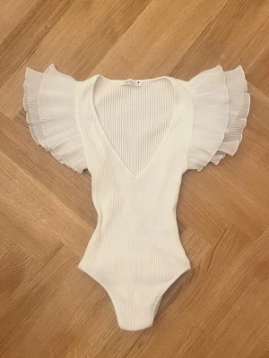 Vit body med volang - Supersnygg vit body med djup V-ringning och breda volangärmar i plisserat tyg💗💗 Helt slutsåld och trendig! Passar ca XS-S. Har en liten lagad defekt, se sista bilden.