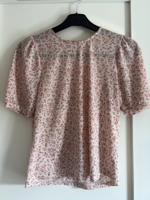 Blommig puffärm blus i ljusrosa - Söt blus med puffärmar och rund halsringning i ljusrosa med små röda blommor. Blusen har en avslappnad passform och är gjord i ett lätt, mjukt tyg som känns skönt mot huden. Perfekt för dig som gillar romantiska och feminina detaljer.