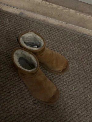 Uggs - Säljer ett par bruna boots med kort skaft och mjukt fårskinnsfoder. Yttermaterialet är mocka och sulan är mönstrad och gjord av gummi. Perfekta för kalla dagar och riktigt sköna att ha på sig. Klassisk design som funkar till många olika stilar.
