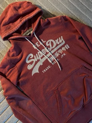 Vinröd hoodie från Superdry - Snygg vinröd hoodie från Superdry med vit logga och japanska tecken på bröstet. Klassisk känguruficka framtill, vita dragsnören och ribbade muddar. Perfekt för en avslappnad och trendig stil.
