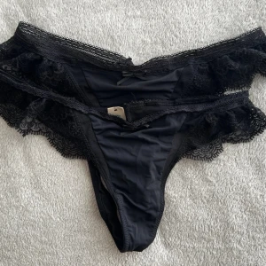 Svarta spets-trosor från Hunkemöller - Säljer ett par snygga svarta trosor från Hunkemöller med breda spetskanter och liten rosett framtill. Trosorna har en klassisk tanga-modell i mjukt, glansigt material och dekorativ spets längs sidorna för en extra feminin touch. 2x par. Ny pris 220kr