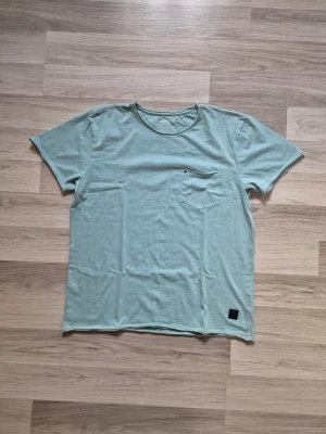 Ljusgrön t-shirt med bröstficka - Snygg ljusgrön t-shirt i bomull med rund halsringning och en liten bröstficka på vänster sida. T-shirten har korta ärmar och en diskret svart patch nertill. Perfekt för en avslappnad stil.