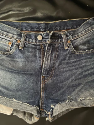 Levi's blå jeansshorts med rå kant - Säljer ett par klassiska blå jeansshorts från Levi's med råa kanter och femficksmodell. Shortsen har hög midja, slitna detaljer och Levi's ikoniska läderpatch bak. Perfekta för sommaren och ger en avslappnad look.