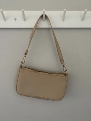 Beige axelremsväska  - Stilren beige handväska med avtagbar axelrem och guldfärgade detaljer. Väskan har en enkel, rektangulär form och dragkedja upptill samt en liten ficka med dragkedja på baksidan. Tillverkad i slätt konstläder som ger en modern look.