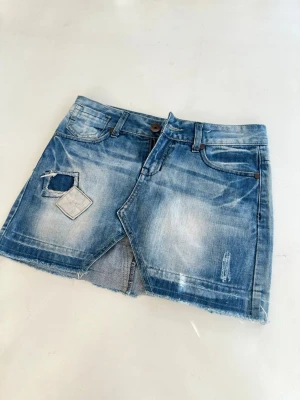Blå jeanskjol med slitningar - Säljer en blå jeanskjol i kort modell med slitna detaljer och rå kant nertill. Kjolen har klassiska fickor fram och bak samt patchade detaljer och en liten slits framtill. Perfekt för dig som gillar en avslappnad och trendig stil. Mått finns på bilderna