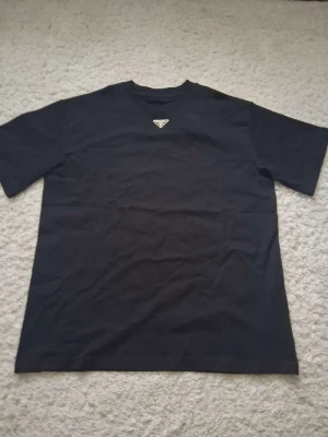 Svart t-shirt från Prada - Snygg svart t-shirt från Prada med det ikoniska metalltriangel-logotypen på bröstet. Klassisk rund halsringning och korta ärmar. Perfekt för dig som gillar stilrena och exklusiva plagg.