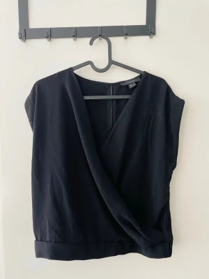 Svart omlottopp  - Snygg svart omlottopp från Atmosphere med djup v-ringning och bred fåll nedtill. Toppen är ärmlös och har en stilren, enkel design som passar till många olika looks. Passar till stl s-m.