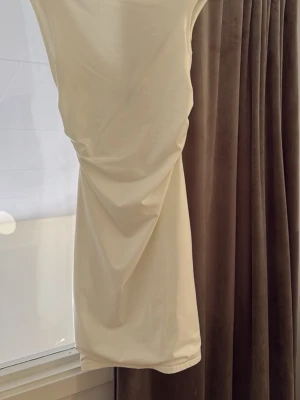 Vit kort klänning/Studentklänning - Säljer en stilren vit kort klänning med breda axelband och snygg drapering i sidan. Klänningen har en djup urringning baktill  och är tillverkad i ett mjukt, stretchigt material som formar sig efter kroppen. Perfekt för dig som gillar minimalistisk och modern stil. Har använt den i några timmar på min student, hade skyddspåse när jag sprang ut så helt fläckfri. 