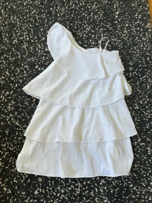 Vit volangklänning från Gina Tricot - Säljer en vit klänning från Gina Tricot med flera lager volanger. Klänningen har oneshoulder-design med ett smalt axelband och är tillverkad i ett lätt tyg, perfekt för varma dagar.
