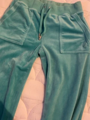 Turkosa mjukisbyxor i velour - Säljer ett par turkosa mjukisbyxor i mjuk velour med bred resår i midjan och dragsko. Byxorna har två stora fickor fram och ett diskret broderat märke på ena fickan. Perfekta för chill dagar och riktigt sköna att ha på sig. Dock fläckar längst ner på ena benet, vet ej om de går bort! annars som nya!!