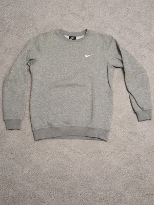 Grå sweatshirt från Nike - Klassisk grå sweatshirt från Nike med rund halsringning och diskret vitt Nike-logga broderad på bröstet. Tröjan har ribbade muddar vid ärmslut och nederkant, perfekt för en avslappnad och sportig stil. Tillverkad i mjukt material för extra komfort.