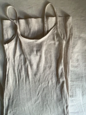 Beige ribbad långklänning - Säljer en beige ribbad långklänning med smala axelband och slits på sidan. Klänningen är ärmlös och har en mjuk, stretchig passform som sitter snyggt på kroppen. Perfekt för varma dagar och enkel att styla med accessoarer.