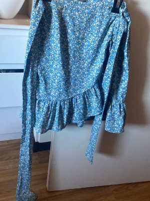 Only Blått blommigt set med volanger - Säljer ett superfint set med omlottopp och matchande kjol i blått med små vita blommor. Båda delarna har volangkant och knytdetaljer i midjan och vid ärmen. Materialet känns lätt och luftigt, perfekt för vår och sommar.  Only 