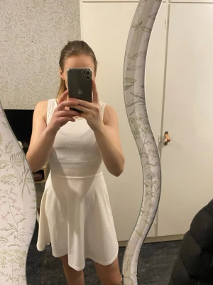 Vit ärmlös kortklänning - Säljer en enkel och snygg vit kortklänning med ärmlös design och rund hals. Klänningen har en lätt utställd kjol och är gjord i ett stretchigt material som ger en bekväm passform. Perfekt för varma dagar eller när du vill känna dig extra fräsch.Fin vit klänning med struktur i tyget. Lite öppen i ryggen.  Inte mycket använd. Kan inte se några fläckar eller skavanker.