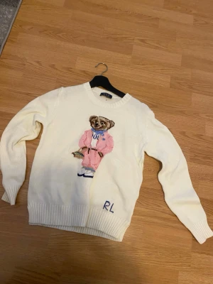 Vit stickad tröja med björn från Polo Ralph Lauren - Vit stickad tröja från Polo Ralph Lauren med ikonisk björn i rosa outfit på bröstet och RL-broderi nedtill. Tröjan har rund halsringning, långa ärmar och ribbstickade muddar. Perfekt för dig som gillar klassisk preppy stil. Inte äkta