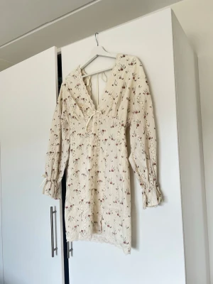 Blommig kortklänning med volang - Söt kortklänning i ljusbeige med små blommor i rött och grönt. Klänningen har v-ringning, volangdetaljer vid halsen och långa ärmar med knyt vid handlederna. Tillverkad i ett lätt bomullstyg, perfekt för varma dagar.