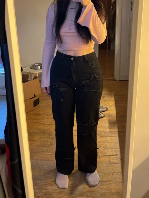 Svarta slitna raka jeans - Säljer ett par svarta jeans med raka ben och slitningar på både fram- och baksida. Jeansen har hög midja, klassiska fem fickor och stängs med knapp och dragkedja. Perfekta för en avslappnad och trendig look.