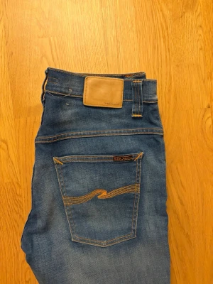 Blå jeans från Nudie Jeans - Säljer ett par klassiska blå jeans från Nudie Jeans med snygga kontrastsömmar och ikonisk våg på bakfickorna. Jeansen har normal passform och är tillverkade i bomull. Perfekta till din vardagsstil!