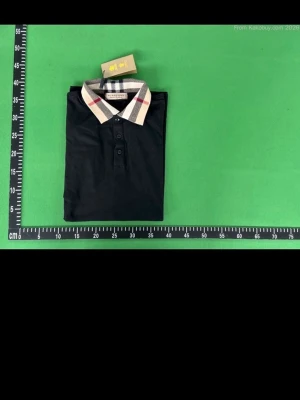 Svart pikétröja med rutigt krage - Svart pikétröja från Burberry med klassisk rutigt mönster på kragen och knappar framtill. Tröjan har korta ärmar och en stilren, tidlös design som passar perfekt till många olika outfits. Tröjan är storlek xxl men passar även xl 