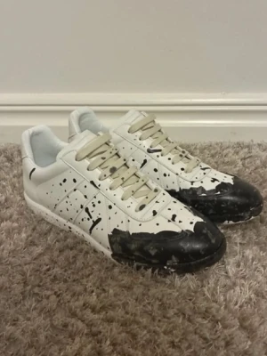 Vita sneakers med svart färgstänk - Unika vita sneakers med svart färgstänk-design på tå och sidor.får inte sammankopplas med Maison margiela för Demma har inge märkningar dessa skor är no branded Skorna har lågt skaft, vita snören och är tillverkade i slätt läder. Perfekta för dig som vill sticka ut med en kreativ och edgy look.