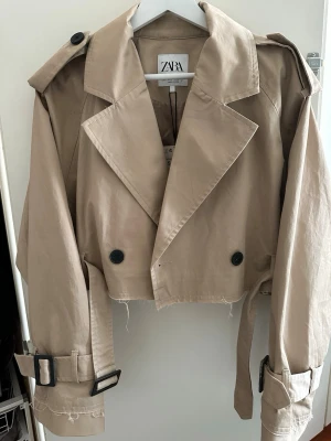 Beige cropped trenchcoat från Zara - Trendig beige cropped trenchcoat från Zara med stora slag, dubbelknäppning och bältesdetaljer vid ärmsluten. Jackan har rå kant nertill för en edgy look och axelklaffar som ger extra attityd. Perfekt för dig som vill ha en modern twist på klassikern.