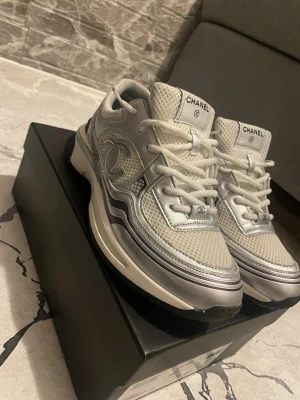 Chanel vita och silver sneakers - Snygga Chanel sneakers i vitt och silver med mesh och läderdetaljer. Klassisk logga på sidan och tungspetsen, samt vita skosnören. Snygg vågig design på sidan och robust sula för extra komfort. Perfekta för dig som vill sticka ut med exklusiva skor. Skor jag jag bara använt några gånger och är typ helt nya. Om du vill se mer bild det är bara att skriva! Priset kan diskuteras!