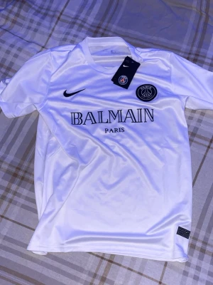 Vit PSG x Balmain tröja Nike - Snygg vit PSG x Balmain Paris tröja från Nike med svart logga och tryck. Klassisk rund hals och korta ärmar, tillverkad i lätt och ventilerande material. Perfekt för dig som gillar fotboll och streetwear.